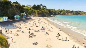 A beach with people sunbathing and swimming, a clear sky, and buildings in the background.