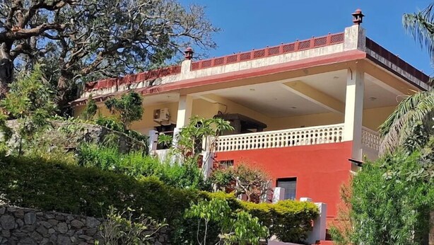 Un edificio de dos plantas con fachada roja y blanca, un balcón y un muro de piedra en primer plano.