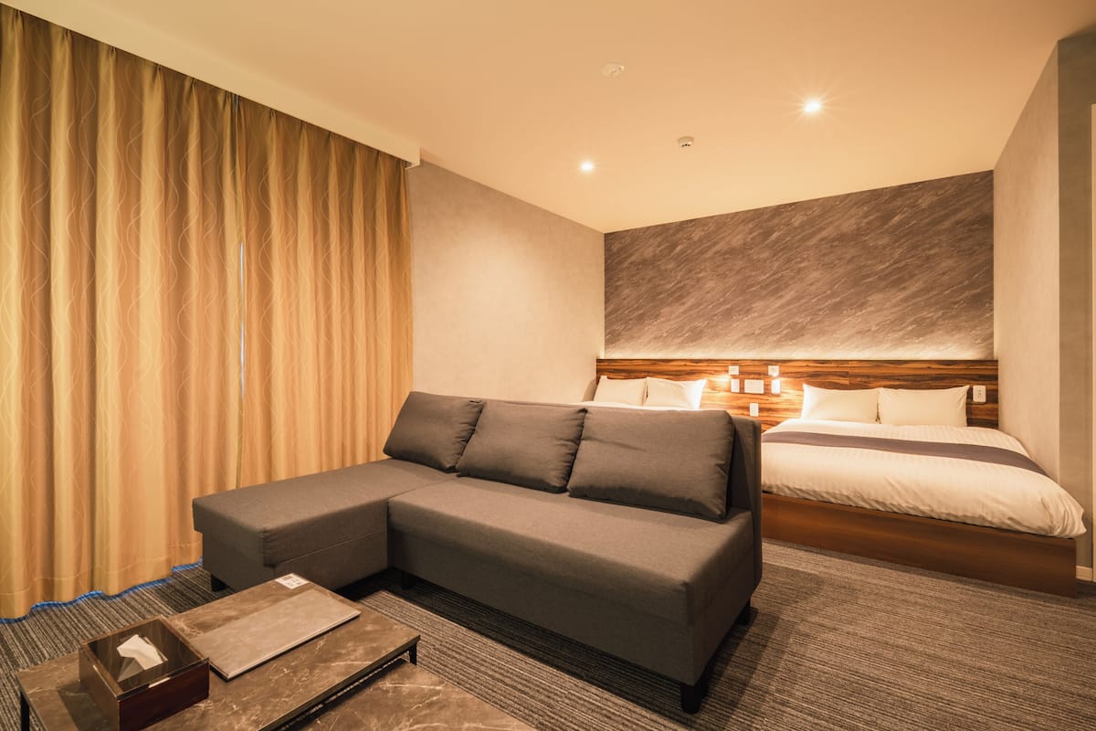 Une chambre d’hôtel moderne avec un canapé d’angle, un lit doté d’une tête de lit en bois et une petite table avec un coffre.