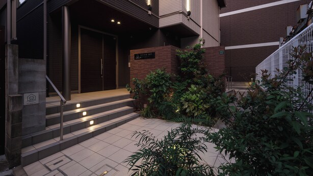 Un bâtiment moderne avec un escalier menant à une porte, entouré de verdure et bordé d’un chemin dallé.