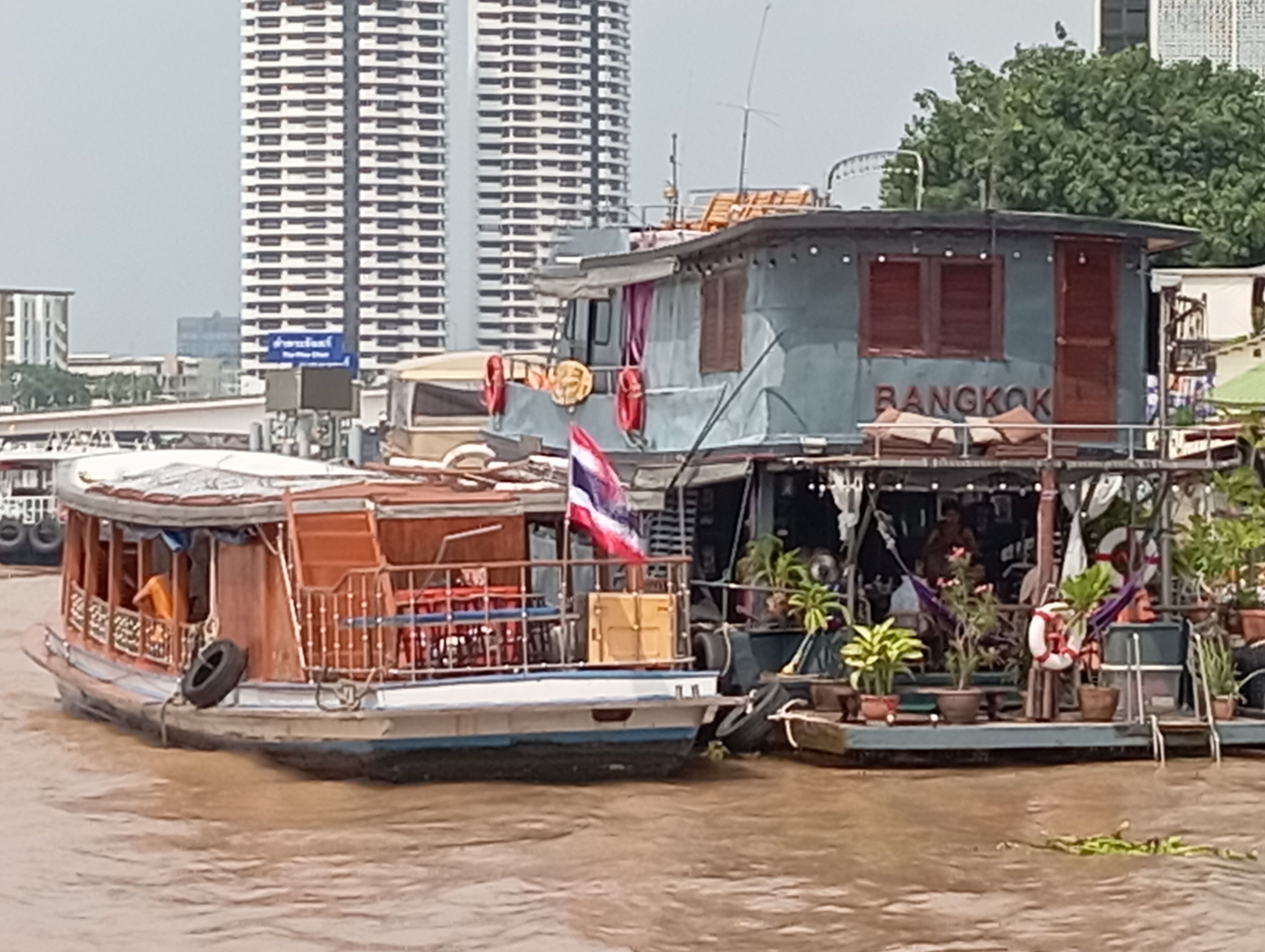 A boat with a covered deck and a flag, docked in a waterway with buildings in the background.
