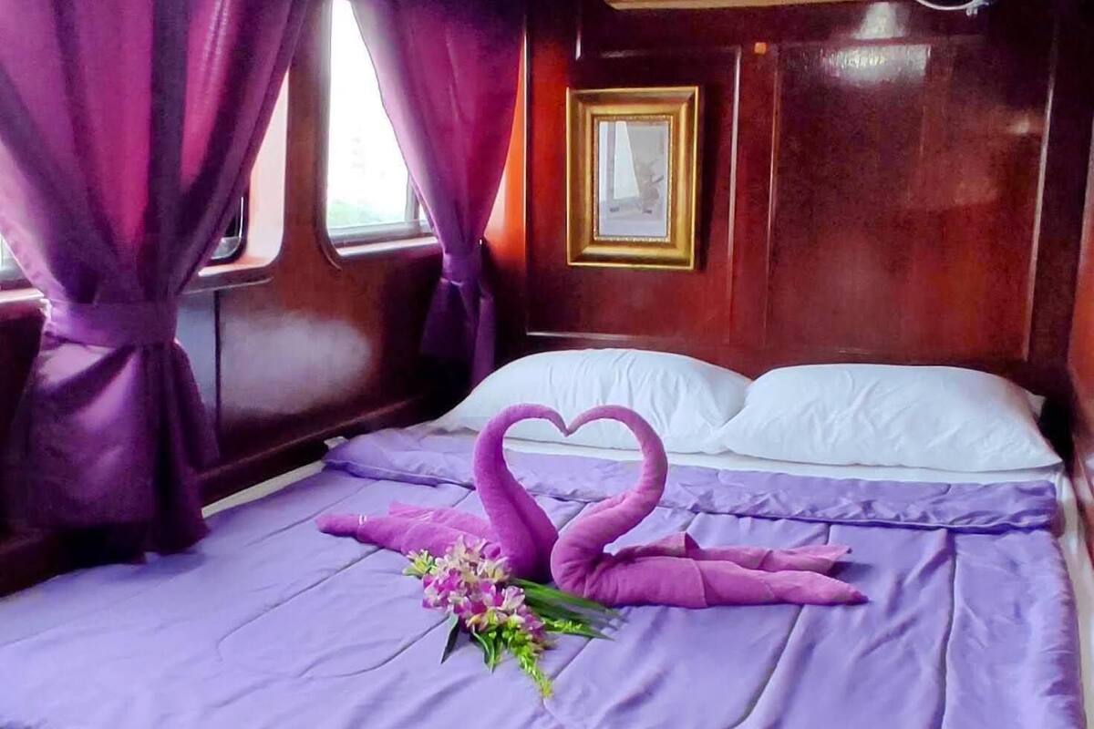 Uma cama com colcha e almofadas roxas, um buquê de flores e uma decoração em formato de coração feita com toalhas.