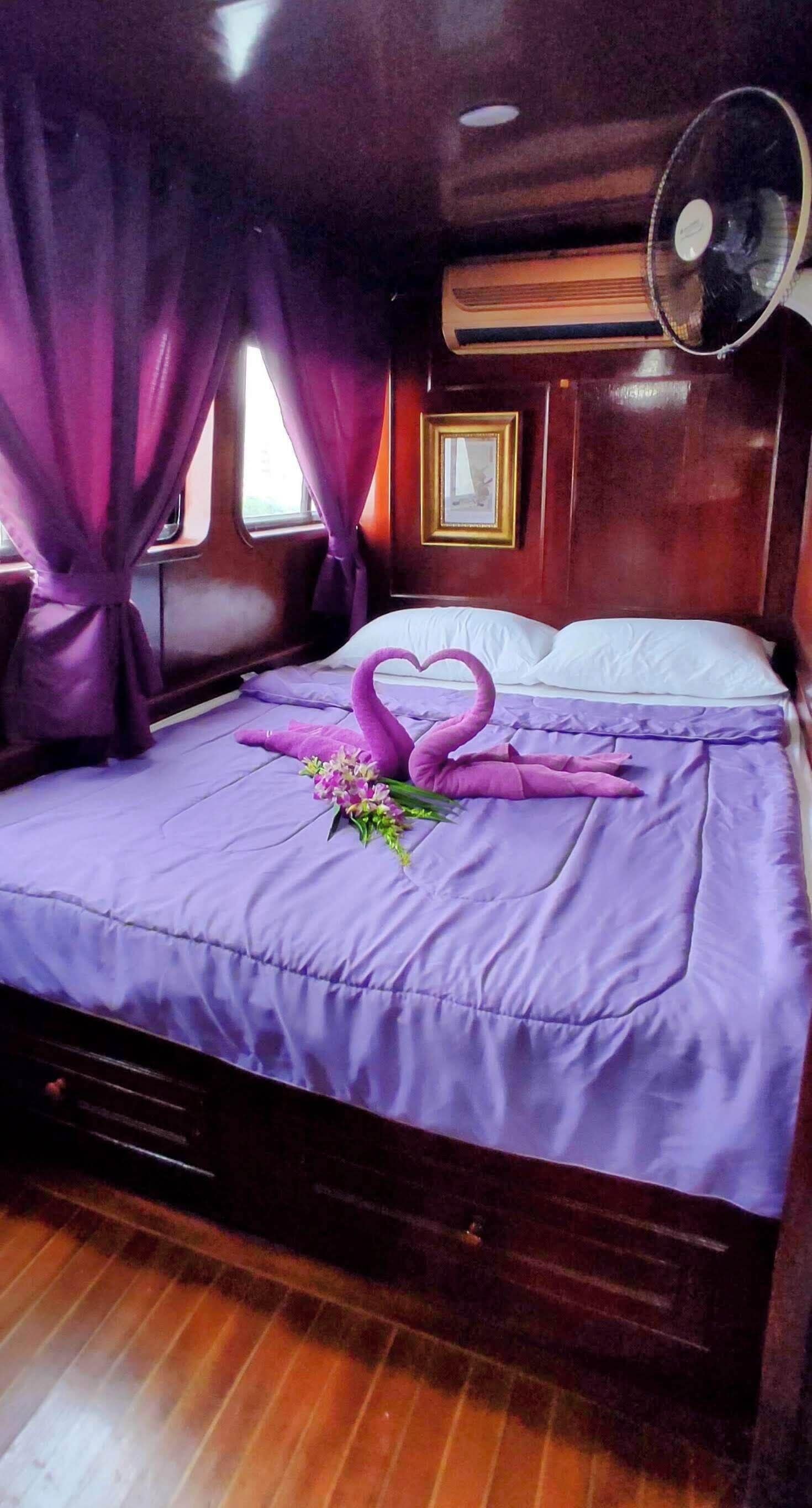 A bed with a purple bedspread, a heart-shaped decoration made of towels, and a bouquet of flowers.