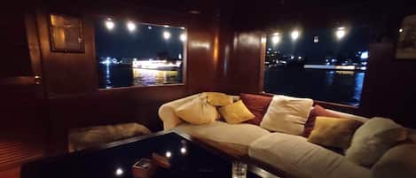 A dimly lit room with a sofa and a glass table, a window overlooking a body of water, and a framed picture on the wall.