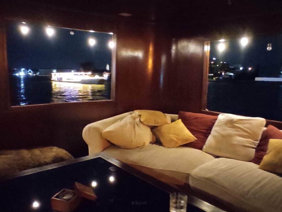 A dimly lit room with a sofa and a glass table, a window overlooking a body of water, and a framed picture on the wall.