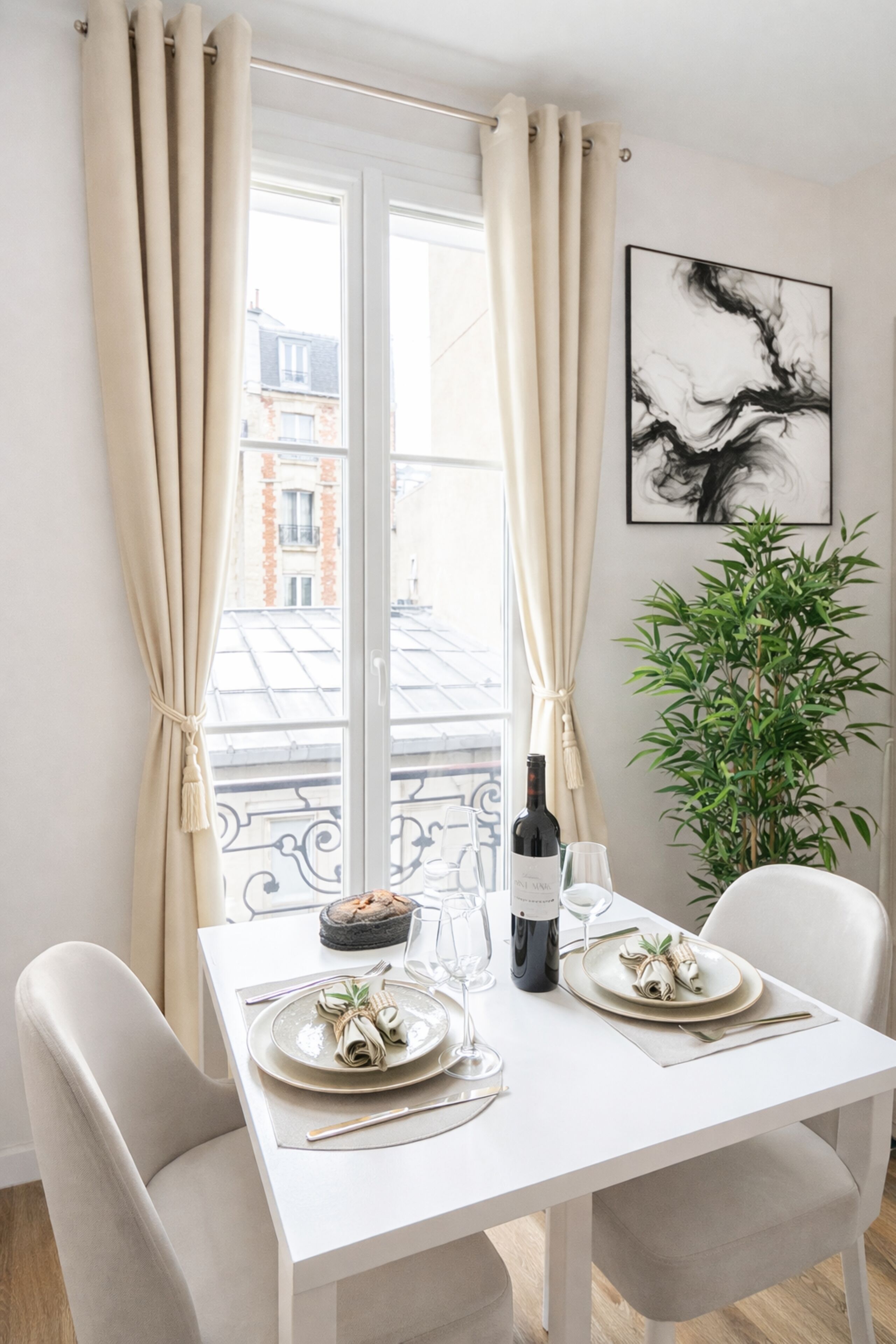 A dining table set with plates, a bottle of wine, and glasses, with a plant and a painting in the background.