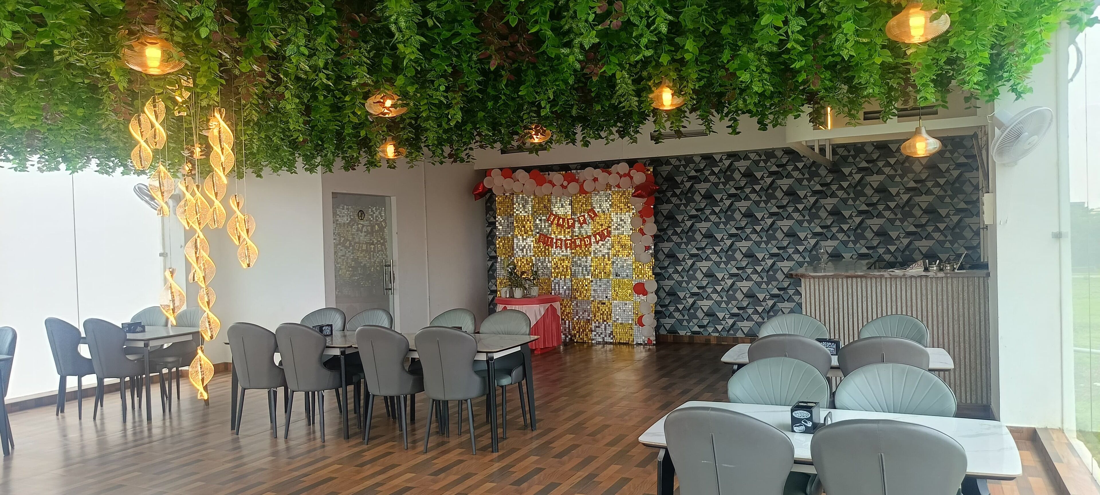 Un restaurante con una decoración moderna, que cuenta con piso de madera, sillas grises y mesas. Se observan luces colgantes y una pared decorativa con un diseño geométrico.
