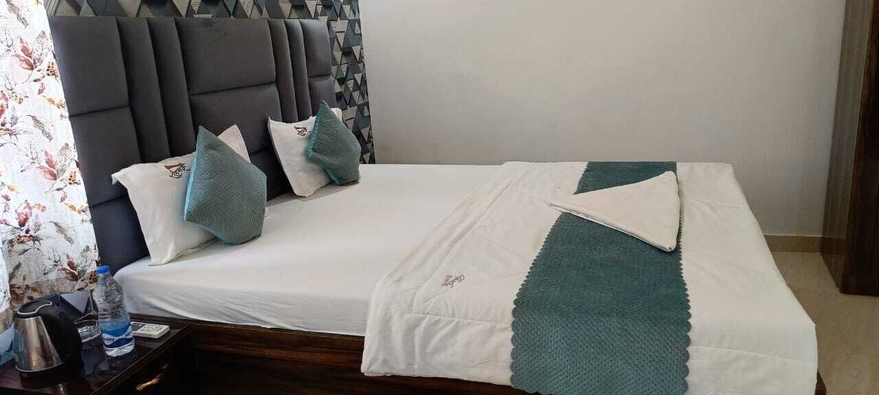 A neatly made bed with a white and teal blanket, a wooden nightstand with a kettle and a bottle, and a headboard with a geometric pattern.