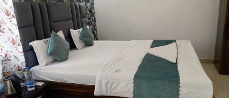 A neatly made bed with a white and teal blanket, a wooden nightstand with a kettle and a bottle, and a headboard with a geometric pattern.