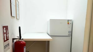 A small room with a white desk, a white refrigerator, a red fire extinguisher, and a sign indicating an emergency exit. - CTA Catania Aeroporto Fontanarossa (Catania)