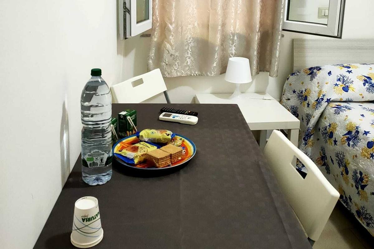 Una habitación pequeña con una cama, una mesa con comida, una botella de agua y una taza.