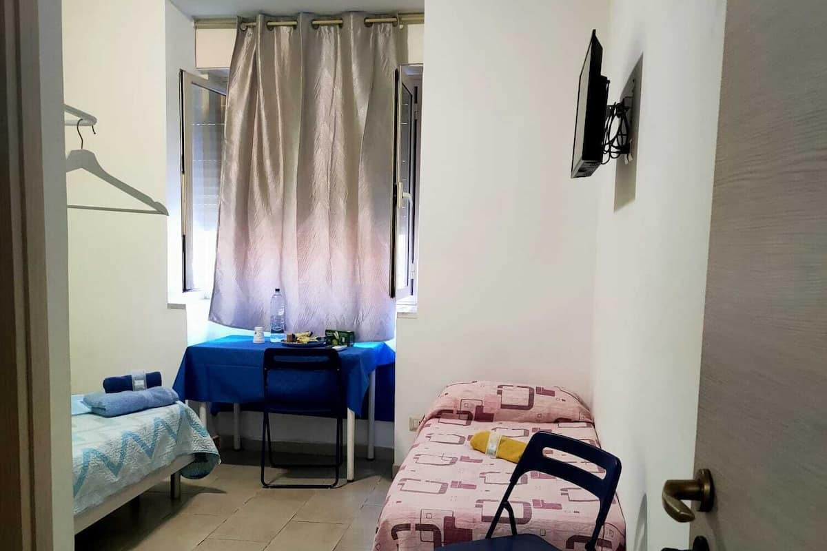 Una habitación pequeña y bien iluminada con una cama, un escritorio con silla, una ventana con cortinas y una puerta.