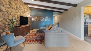 A living room with a stone wall, a grey sofa, a blue accent wall, and a kitchen area visible through an open doorway.