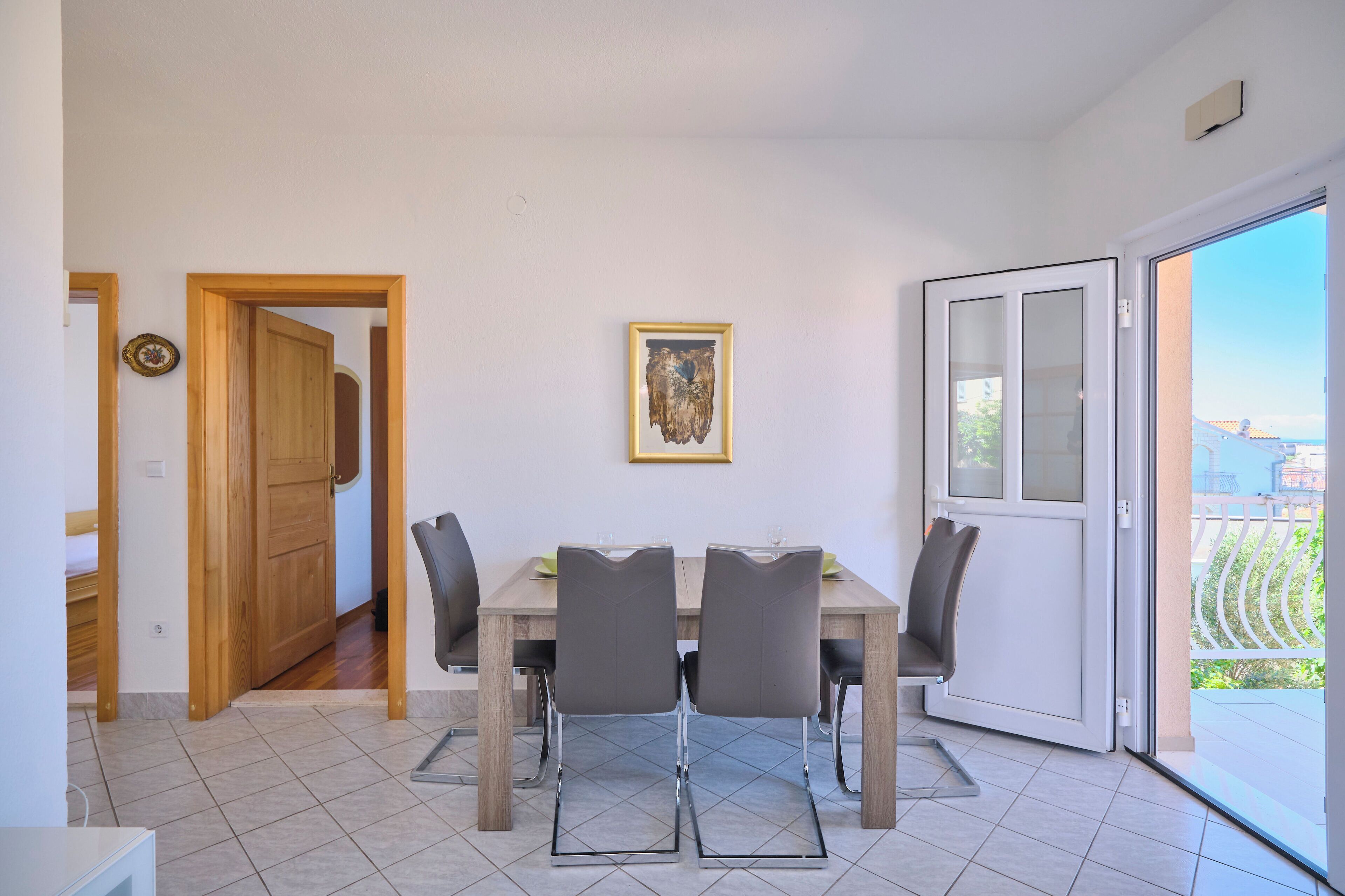 A dining area with a wooden table and four chairs, a framed artwork on the wall, and a door leading to a balcony with a view.