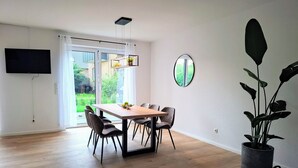 A modern dining area with a wooden table and chairs, a flat-screen TV mounted on the wall, and a large potted plant. - Vacation home Wolke7, with garden, 250 meters to the lake (Lembruch)