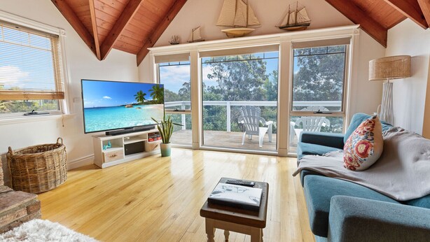 Amplio salón con televisor grande, piso de madera, balcón con sillas y vista al océano.