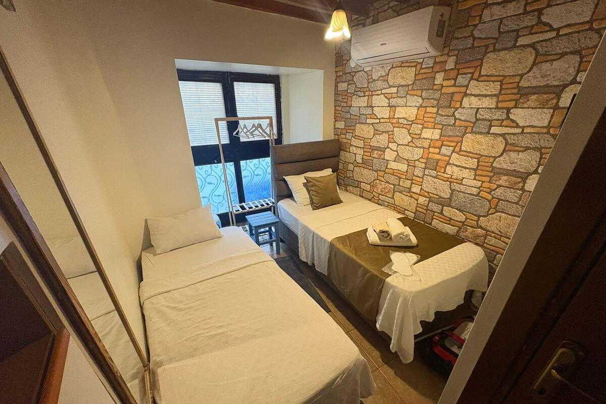 A small, cozy room with two beds, a stone wall, and a window with blinds.