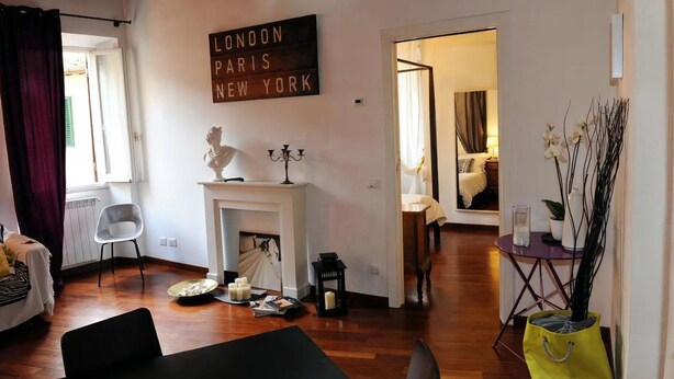 Un salon avec une cheminée, une enseigne indiquant « LONDRES PARIS NEW YORK », et un coin repas avec une table et des chaises.