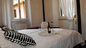 Une chambre d’hôtel avec un lit, un plateau avec des verres à vin et une bouteille, et une fenêtre avec des rideaux.