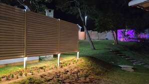 A wooden fence with a metal base, a grassy area with a small tree, and a purple-lit background. - Suite with air conditioning in Praia Seca - Araruama. (Araruama)
