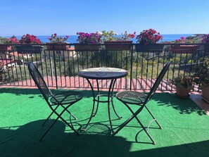 Ein Balkon mit künstlichem Rasen, zwei Metallstühlen, einem runden Tisch mit Mosaikplatte und Kübelpflanzen mit rosa Blüten.