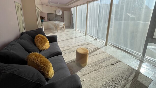 A modern living room with a grey sofa, two fluffy cushions, a round wooden stool, and a large window with sheer curtains.