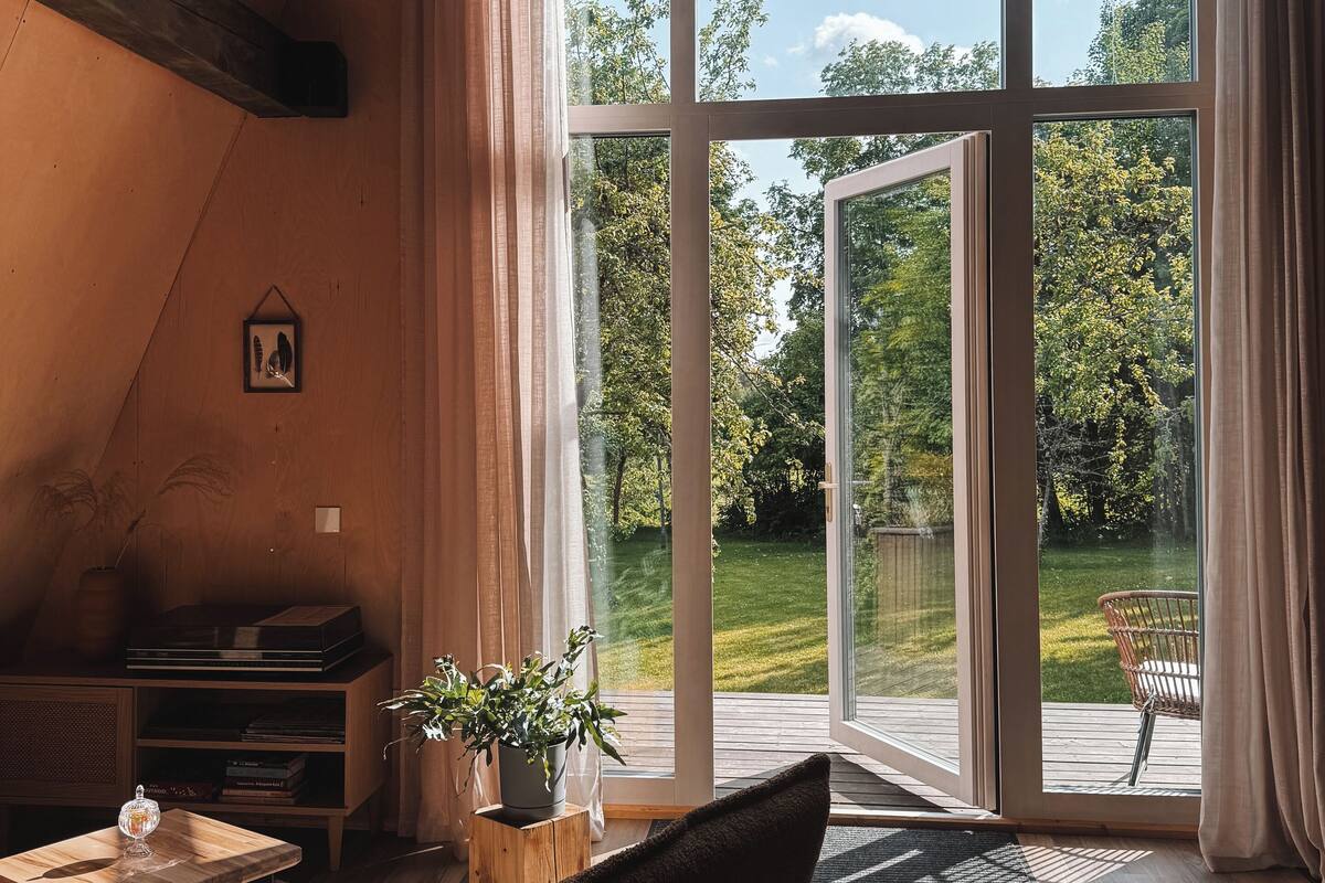 Ein geräumiger Raum mit einem großen Schiebeglasfenster, das zu einem Außenbereich führt, Holzbalken an der Decke, einer Kristallleuchter und einer gemütlichen Sitzecke mit Teppich und Pflanze.