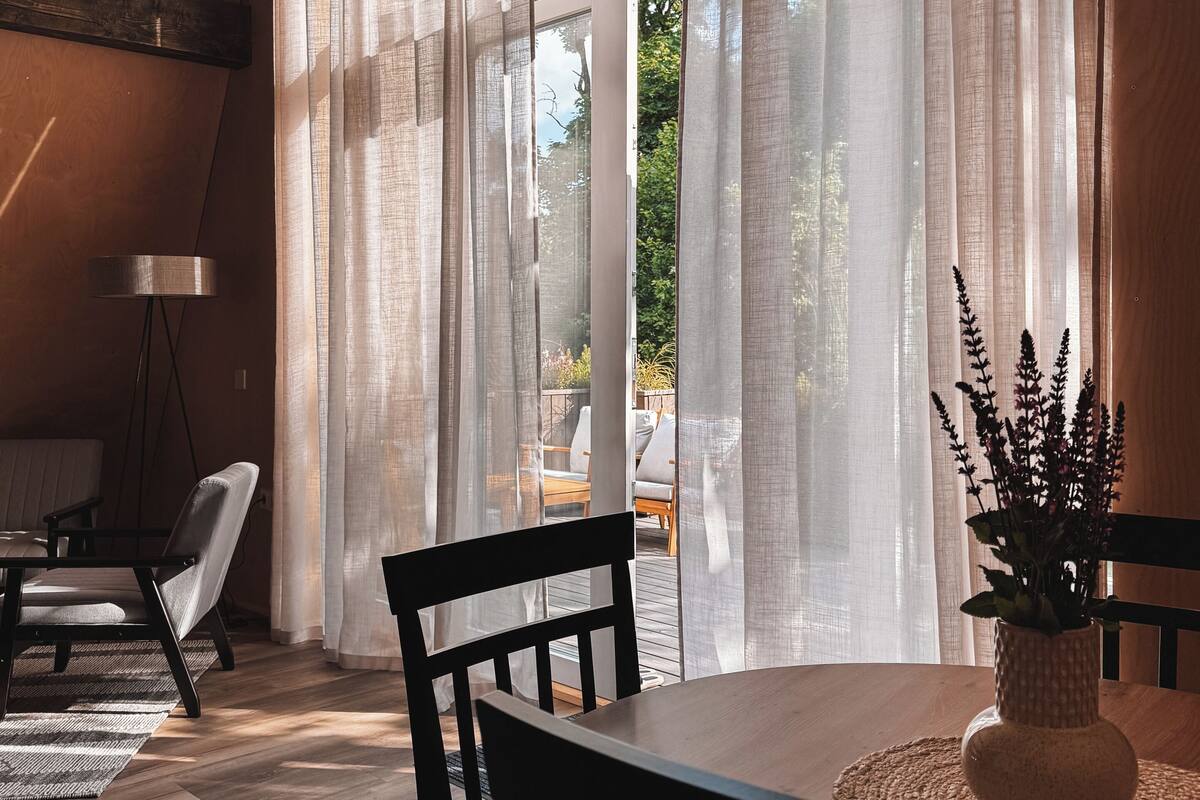 Ein modernes Interieur mit Holzboden, einem Esstisch mit Stühlen, einer Stehlampe und transparenten Vorhängen.