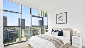 A modern bedroom with a large window, a bed with white bedding, a headboard, bedside tables with lamps, and a framed picture on the wall. - Waterloo Serenity Suite (Waterloo)