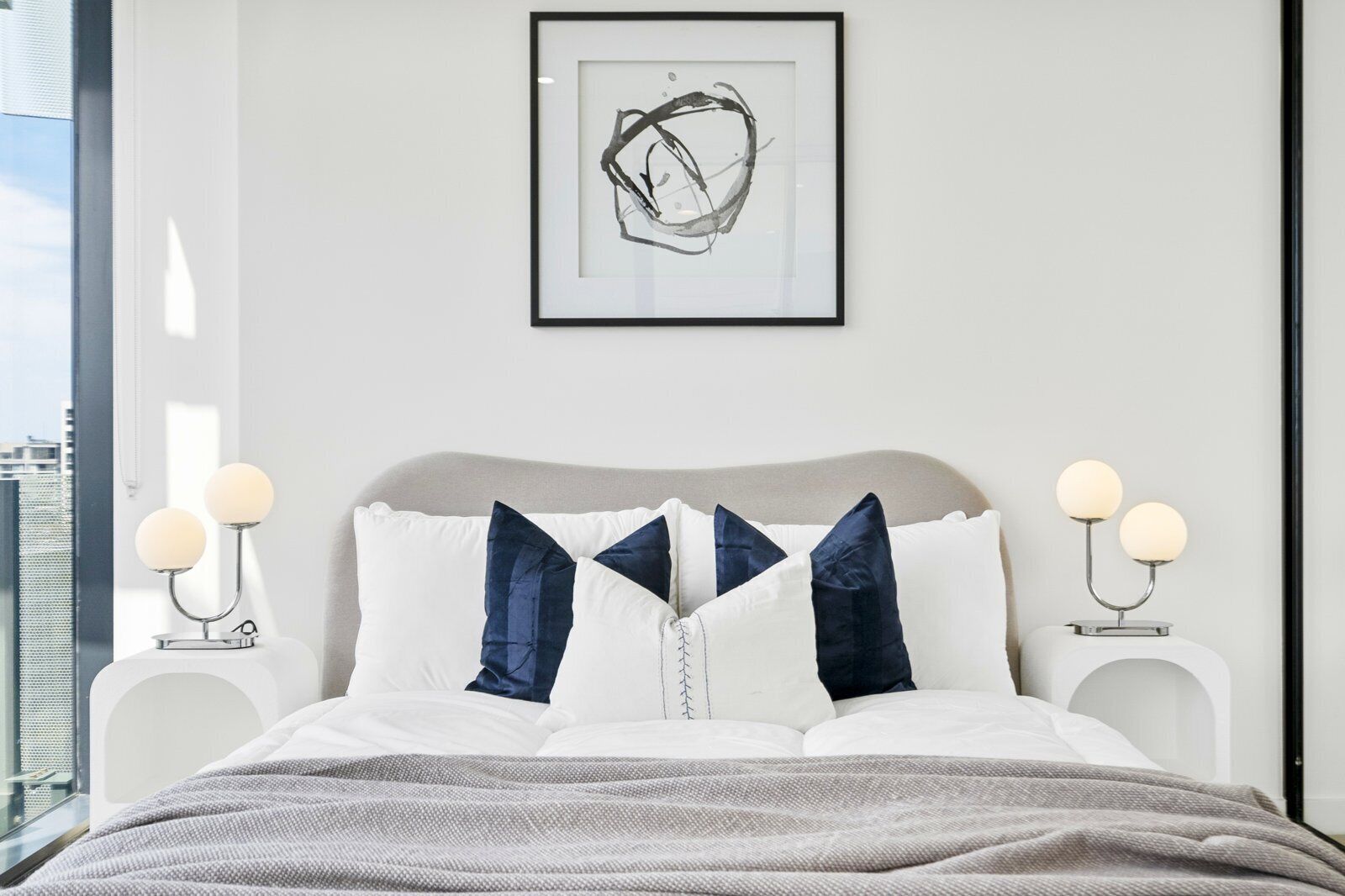 A modern bedroom with a bed, pillows, bedside tables, and a framed abstract artwork.