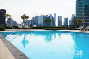 Piscina com espreguiçadeiras e guarda-sóis, rodeada por edifícios modernos e guindastes de construção.