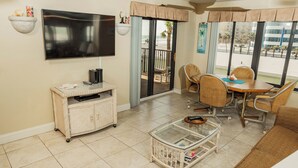 A living room with a flat-screen TV, wicker furniture, a glass coffee table, and a view of the beach through the sliding glass door. - Ocean Delight 2/2 Sandpoint Condo Great OceanViews Pool, Great Rates (Daytona Beach)