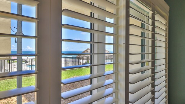 Vista a través de una ventana con persianas horizontales, que da a una playa con una estructura de techo de paja y un área de piscina.