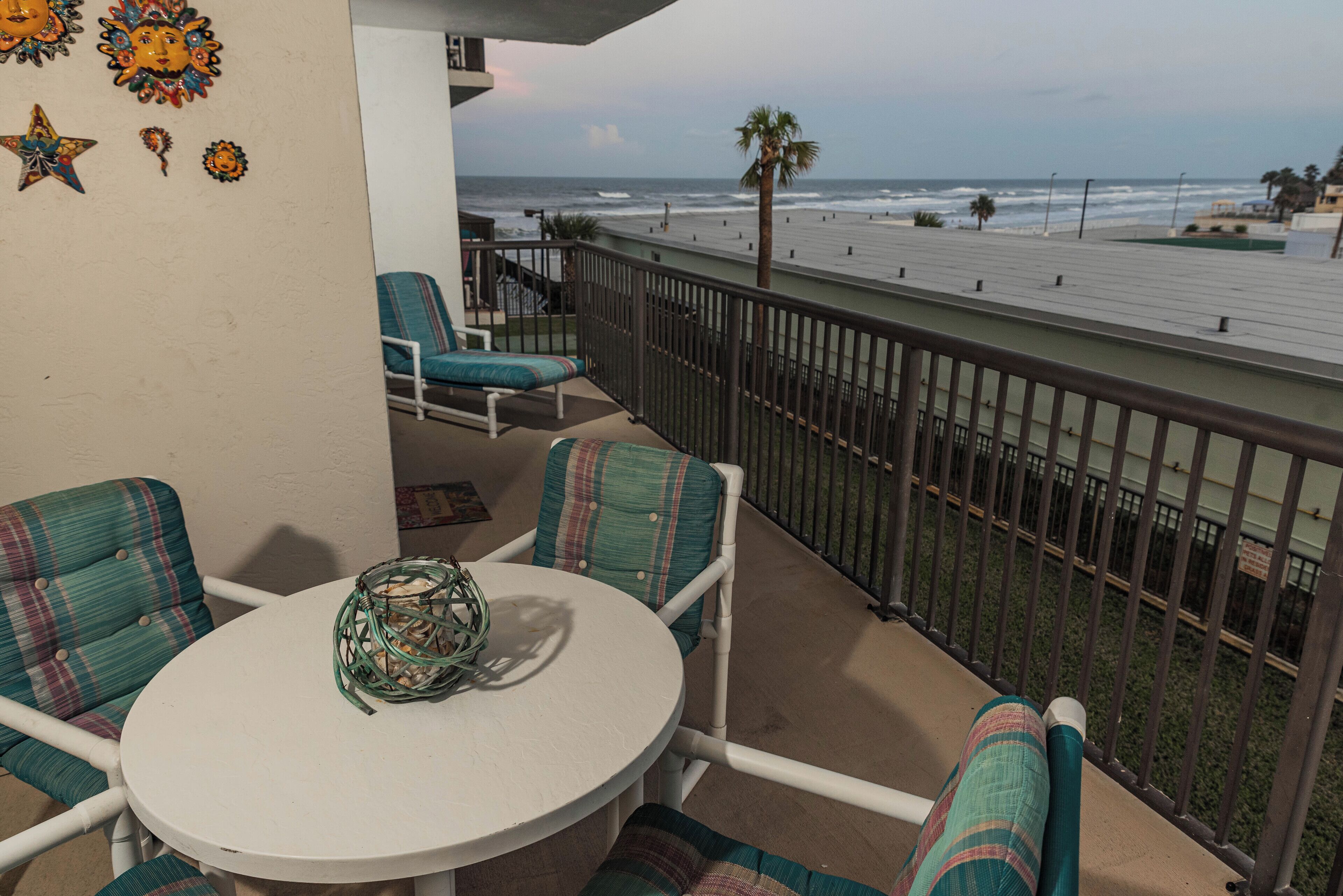 A balcony with a round table and chairs, overlooking a beach with palm trees and a railing.