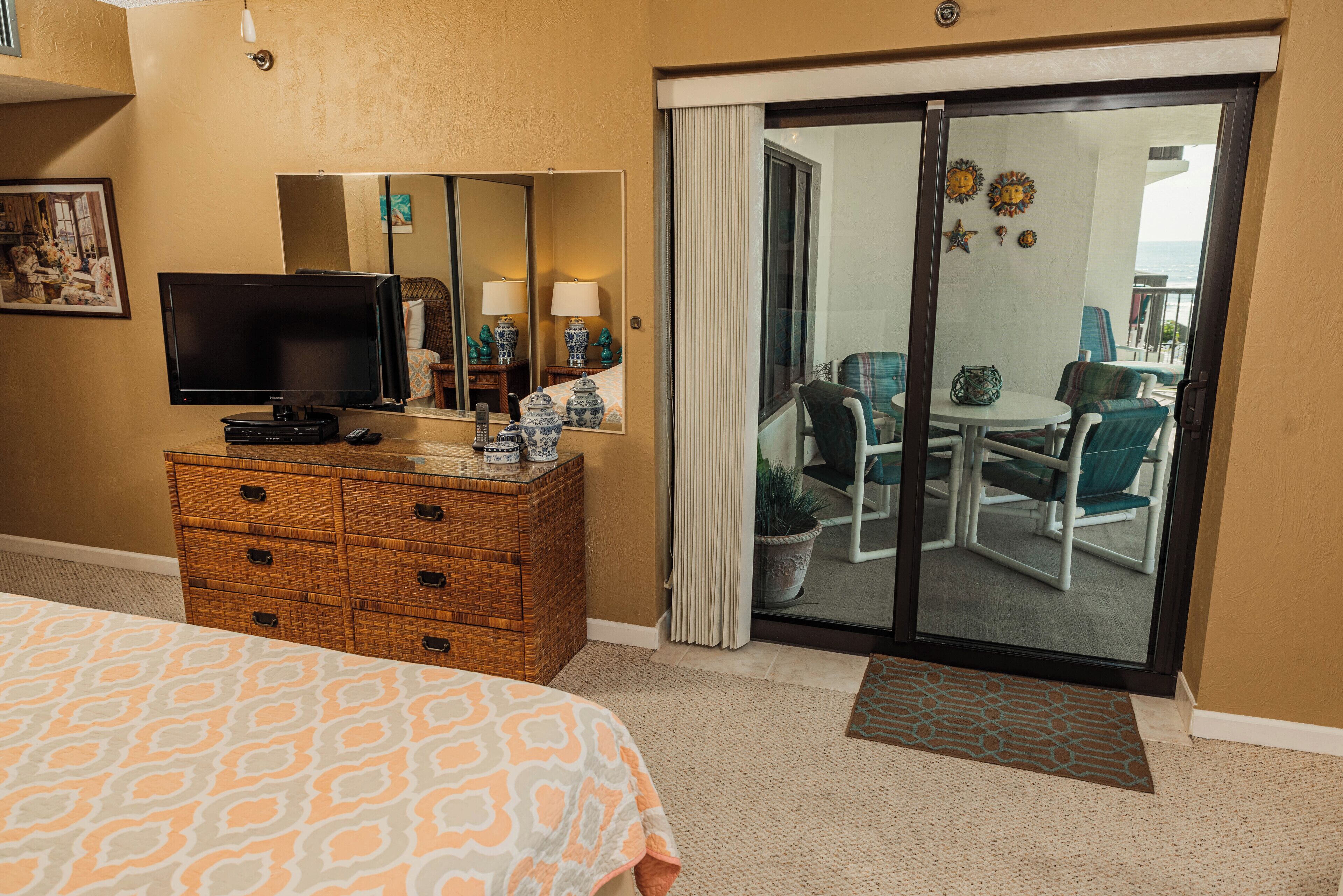 A hotel room with a bed, a dresser, a television, a sliding glass door leading to a balcony with chairs and a table.