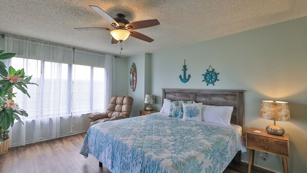 A bedroom with a bed, a chair, a nightstand, a lamp, a ceiling fan, and a window with sheer curtains.