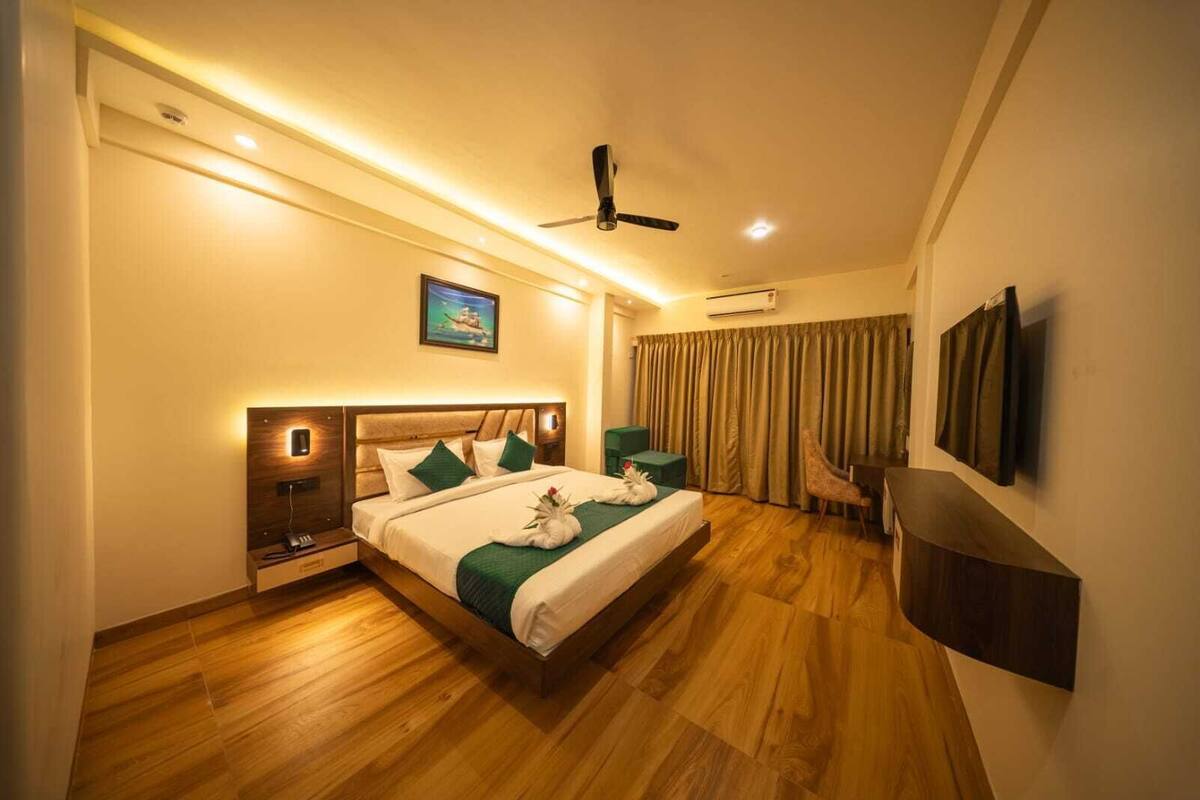 Habitación de hotel moderna con piso de madera, cama grande con cubrecama verde y blanco, ventilador de techo, televisor montado y un cuadro en la pared.