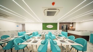 A modern restaurant with teal chairs and white tables, a green wall with a logo, and a marble floor. - Hotel Navil Exotica (Vasco da Gama)