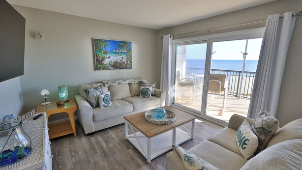 A cozy living room with a sofa, coffee table, and a painting of a tropical scene. - Coastal Charm, 1stFL Stunning Oceanfront 2BR Condo, Lg Balcony, Pool (Daytona Beach)