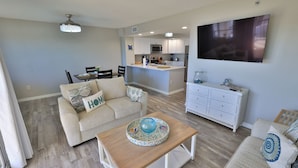 A modern living room with a beige sofa, a coffee table, and a TV. - Coastal Charm, 1stFL Stunning Oceanfront 2BR Condo, Lg Balcony, Pool (Daytona Beach)