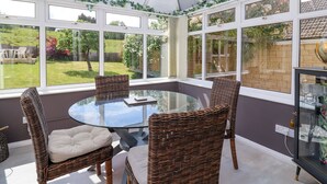 A conservatory with a glass table surrounded by wicker chairs, large windows, and a view of a garden.
