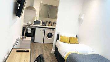 Une chambre d’hôtel compacte comprenant un lit, un petit bureau, une chaise, un lave-linge et une kitchenette.
