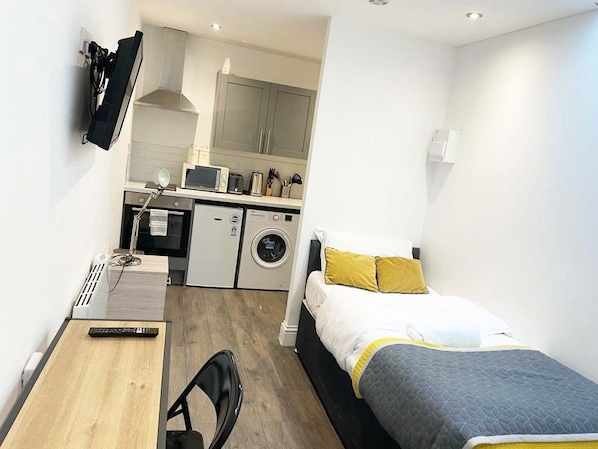 Ein kleines Hotelzimmer mit Bett, Schreibtisch, Stuhl, Waschmaschine und Küchenzeile.