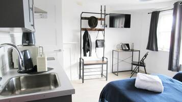 A compact hotel room with a kitchenette, a bed with a blue blanket, a desk with a laptop, and a chair.