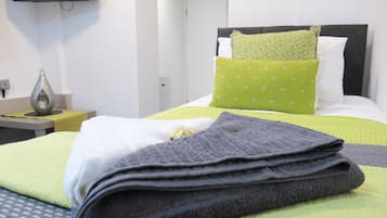 A neatly made bed with a green and white pillow, a folded towel, and a bedside table with a lamp.