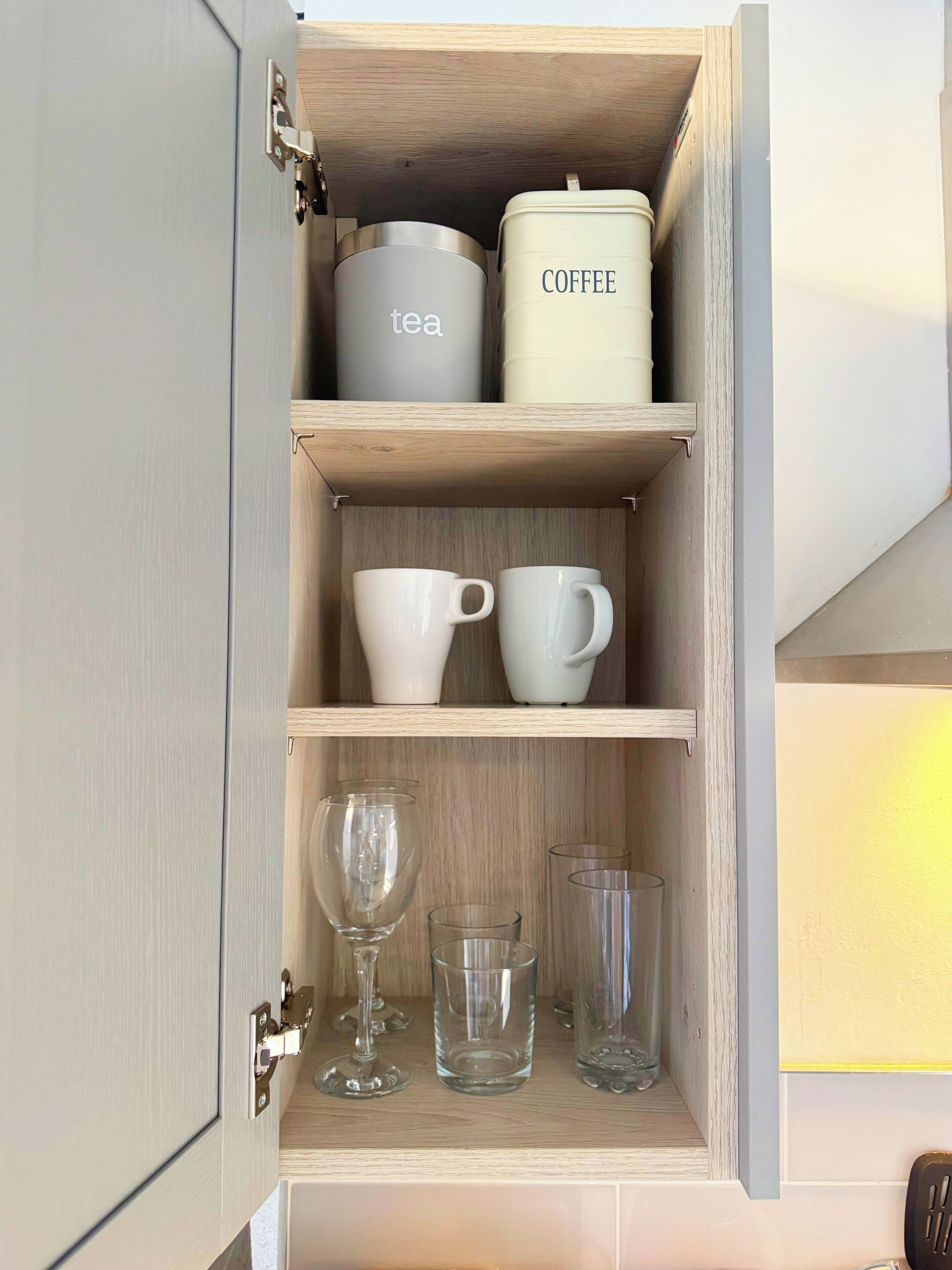 Een keukenkastje met theezeefjes en koffiecontainers, witte mokken en glazen.