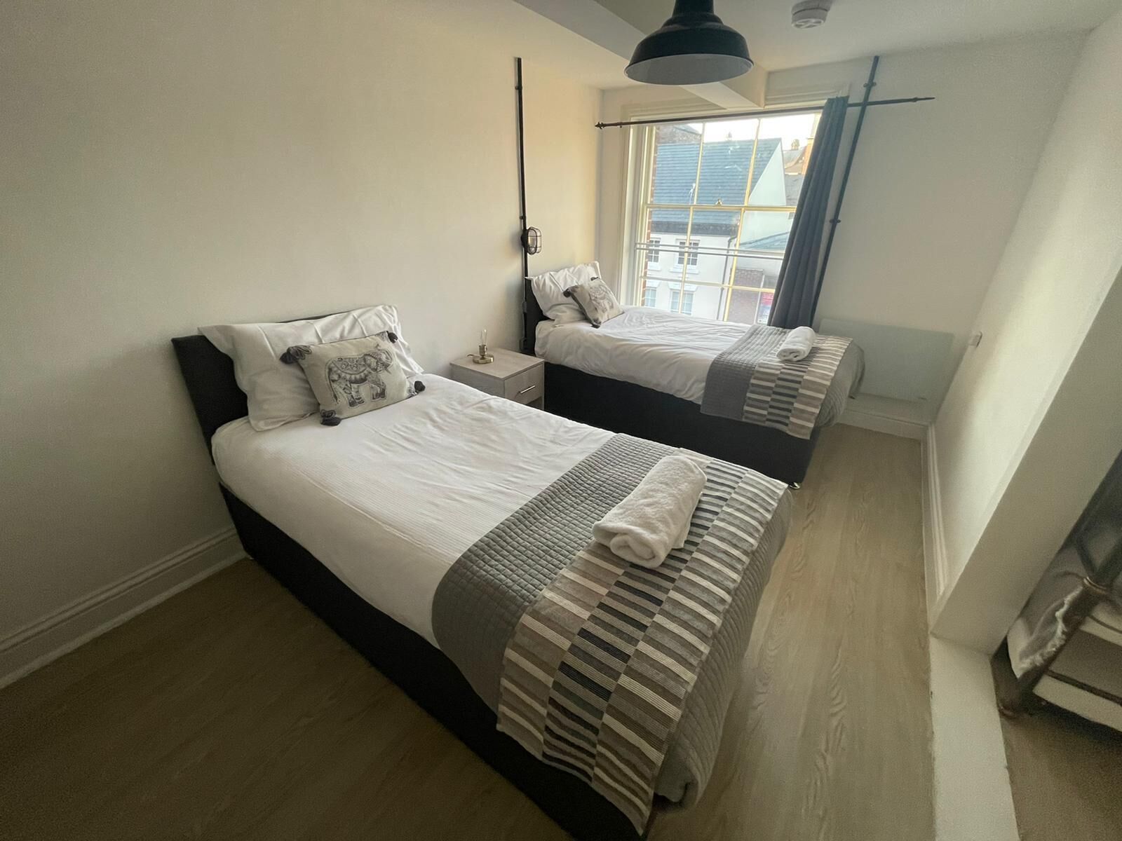 A bedroom with two beds, a nightstand, a window with a view of buildings, and a hanging light fixture.