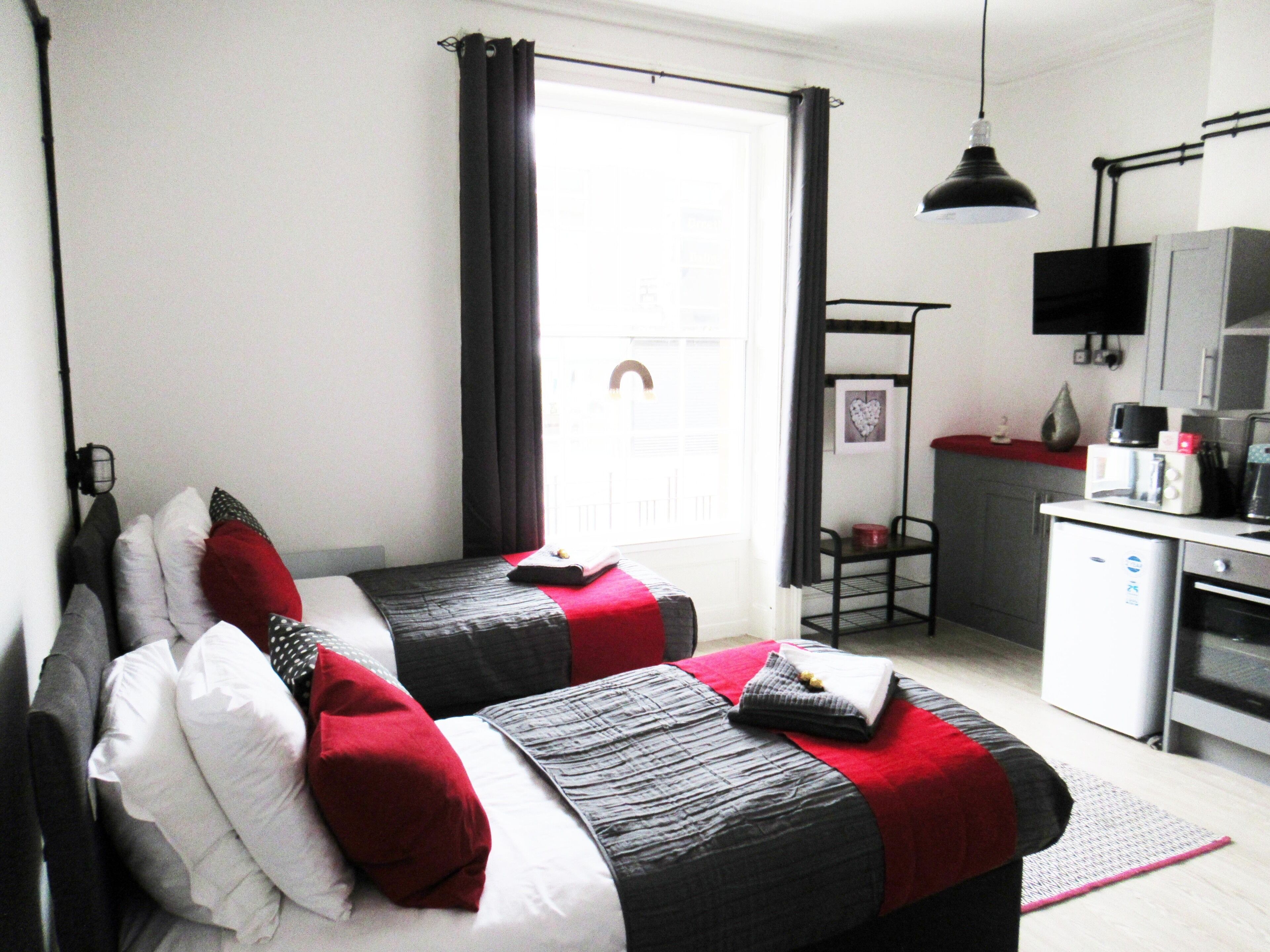Habitación de hotel compacta con dos camas, una kitchenette, un pequeño comedor y un amplio ventanal con cortinas.