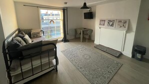 A modern living room with a black metal bed, a large window with a view of buildings, a small table with chairs, a TV on the wall, and a patterned rug. - Queen Street Apartments (Wolverhampton)