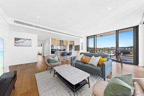 A modern living room with a sofa, chairs, and a dining table. A kitchen is visible in the background, and large windows offer a view of the city.